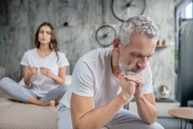 Collect my thoughts. Unhappy pensive adult gray-haired man sitting on bed and young wife at distance