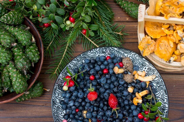 Collected edible orange chanterelle mushrooms on a wooden plate with cranberries blueberries and strawberries Wooden plate with mushrooms green spruce branches and fir cones