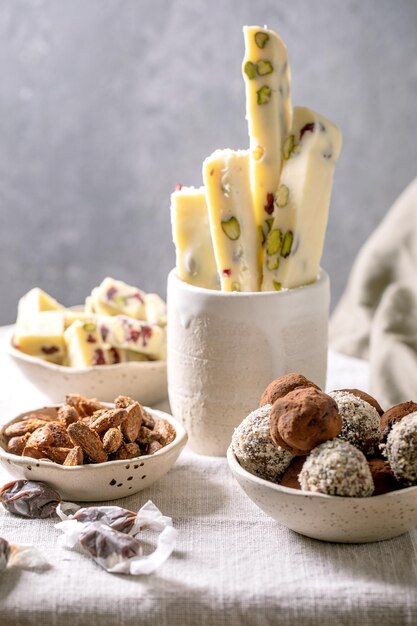 Collection of homemade sweet almond nuts, dark chocolate\
truffle, white chocolate candy bar in bowls on white linen table\
cloth. handmade food sweet gifts for any holidays. sweet table