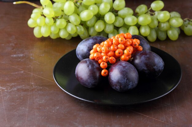 Collection of raw and organic autumn fruits from the garden closeup Ripe juicy grapes plum and rowan on a black plate on a dark background