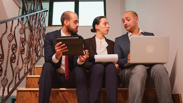 Collega's bespreken plannen die werken aan een moeilijke deadline zittend op een trap in een kantoorgebouw met behulp van laptop en tablet die rapporten analyseert. Groep bedrijfsleider en office executive op trappen.