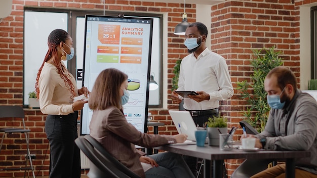 Collega's die bedrijfsstrategie presenteren met analyse op monitor tijdens coronaviruspandemie. Mensen met gezichtsmasker plannen ontwikkelingsproject voor bedrijfsgroei op briefingconferentie.