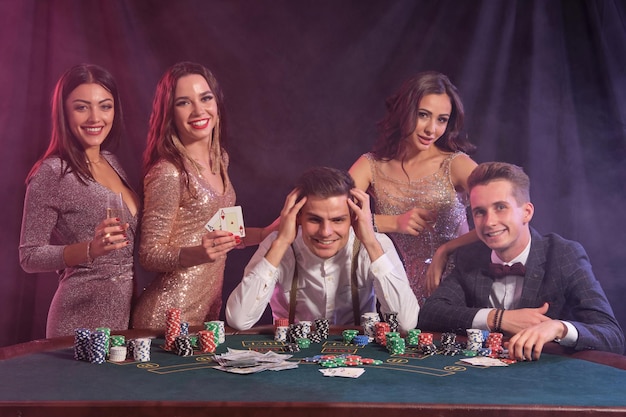 Collega's die poker spelen in het casino aan de tafel met stapels chips geldkaarten erop Vreugde winnen drinken champagne glimlachen Zwarte rook achtergrond kleurrijke achtergrondverlichting Gokken close-up