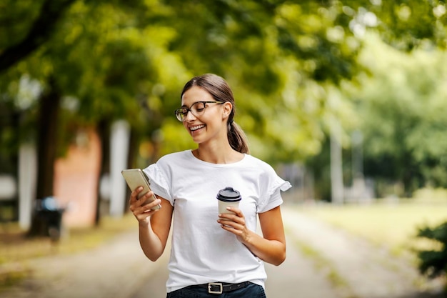 女子大生がキャンパスを歩いていて、電話で重要なメッセージを受け取っている 授業の合間にコーヒーブレイクをしていて、使い捨てのコーヒーカップを持っている
