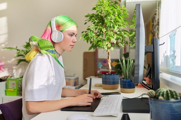 대학생, 헤드폰을 끼고 집에 있는 매력적인 10대 소녀, 그래픽 태블릿으로 컴퓨터에서 공부하고 애완용 녹색 앵무새가 어깨에 앉아 있습니다.