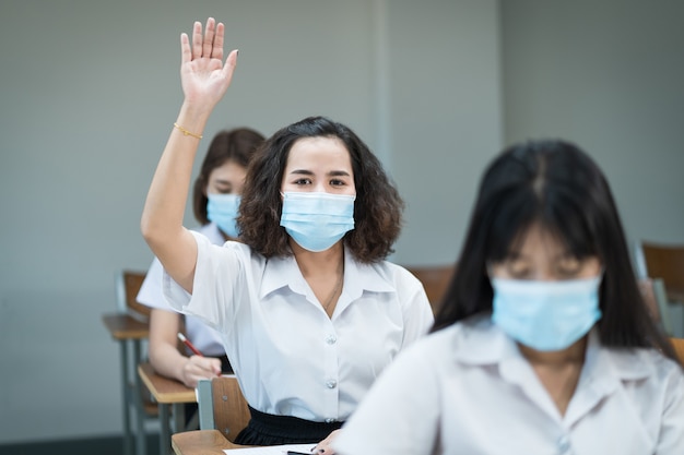 College students wear face mask study in classroom and raise hand to ask teacher during coronavirus pandemic. Selective focus portrait of university students study in the classroom.