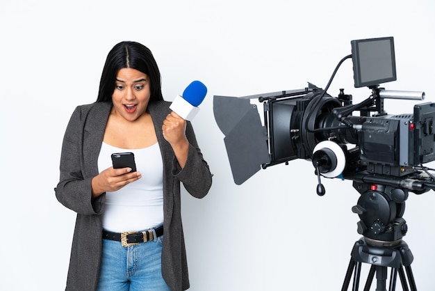 Colombian reporter woman holding a microphone and reporting news on white wall surprised and sending a message