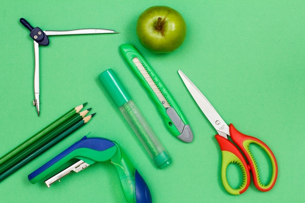 Color pencils, compass, stapler, felt pen, paper knife, apple and scissors on green background. Top view. Back to school concept. School supplies