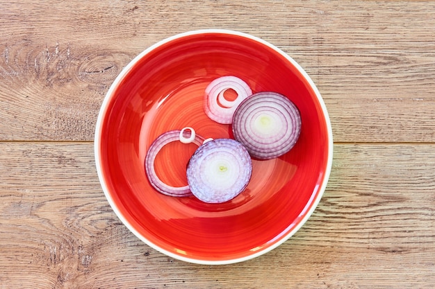 Фото Цветной натюрморт - нарезанная кольцами луковица сладкого фиолетового лука на красной тарелке на деревянной столешнице