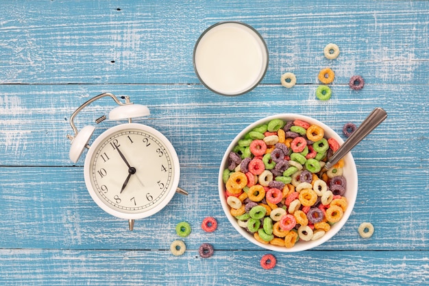 Colored breakfast cereals in a bowl white alarm clock and glass of milk on a blue wooden background healthy breakfast concept for kids top view