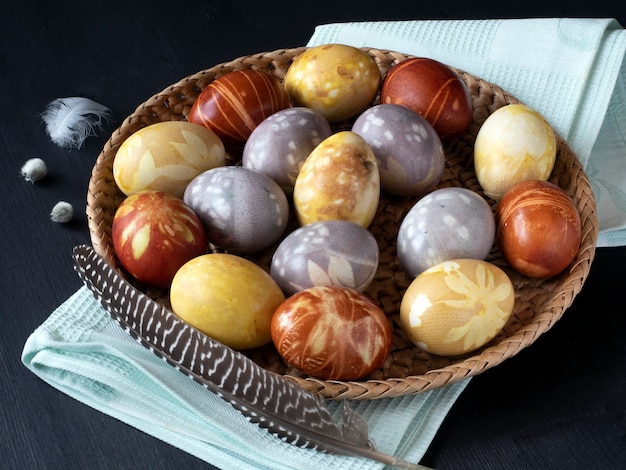 Цветные пасхальные яйца кладут на плетеный поднос на полотенце и на черный деревянный стол.