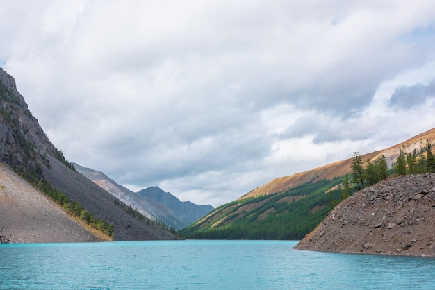 写真 色とりどりの秋の風景森の丘の間にある大きな青青の山の湖変動する天候のい岩のシルエットに反して雲の空の下の日光に照らされた谷の青いアルプス湖に輝く波紋