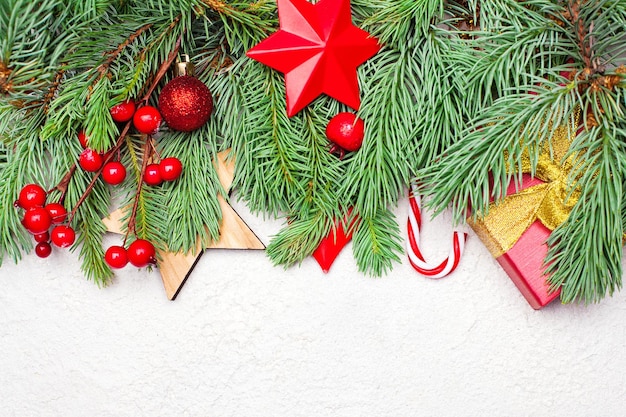 Colorful background with Christmas tree branch, red holly berries, gift, candy and baubles. Christmas composition, top view flat lay with copy space on white stucco plaster texture