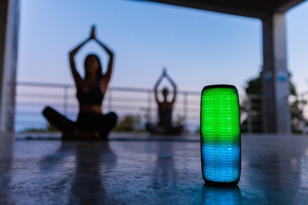 Colorful backlight wireless speaker on foreground of company of friends doing yoga listening to music in dusk