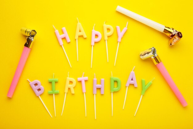 Colorful candles in letters saying Happy Birthday with party foil whistle on yellow background