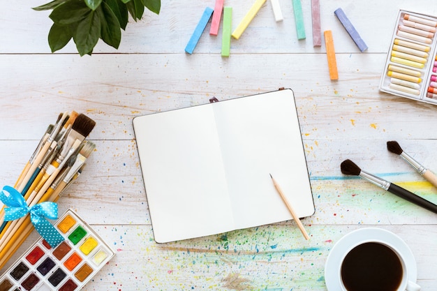 Colorful creative table with blank notebook for sketches and paints, top view