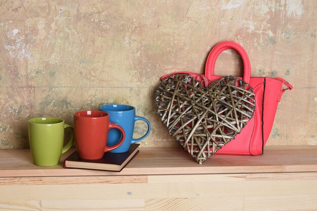 Colorful cups on book and big wooden brown heart with pink ladies handbag on pastel textured and wooden background, copy space