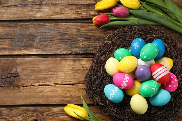 Colorful Easter eggs in decorative nest and tulips on wooden background flat lay Space for text