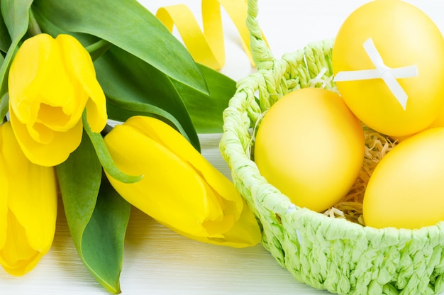 Colorful Easter eggs in green basket and bouquet of tulips on white wooden background. Easter holiday concept.