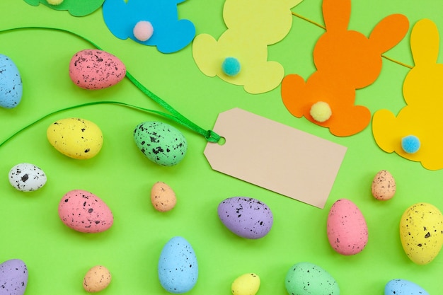 Colorful Easter eggs and handmade bunny easter garland with empty tag on green background. Top view, flat lay. Happy Easter concept.