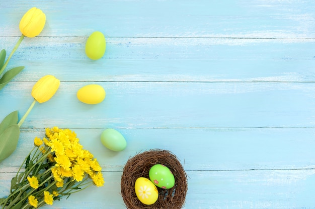 Colorful Easter eggs in nest with flowers on blue wooden background.