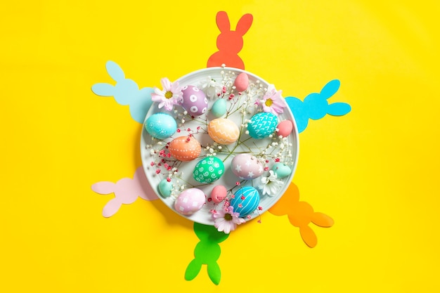 Colorful easter eggs with flowers on ceramic plate with paper bunnies on yellow background