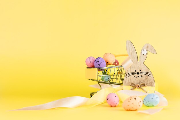 Colorful easter quail eggs in shopping cart and wooden bunny on yellow background Copy space Minimal Easter concept