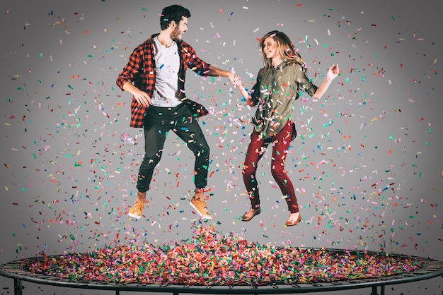 다채로운 재미. 주위에 색종이 조각과 함께 트램폴린에서 점프하는 동안 손을 잡고 있는 아름다운 젊은 쾌활한 커플의 공중 샷