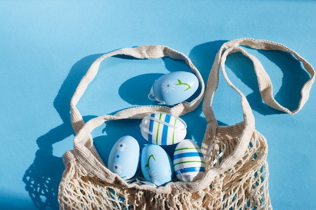 Colorful handmade painted easter eggs in a string bag on blue background with hard shadows