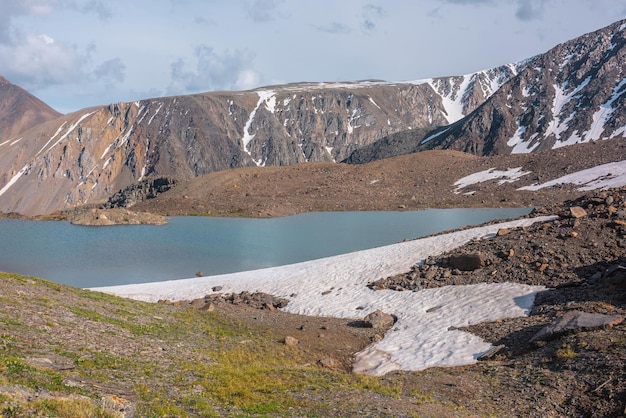 Colorful landscape with beautiful lake in sunlit mountain valley near high mountain range under cloudy sky in changeable weather Awesome scenery with alpine lake among rocks and snow in sunlight