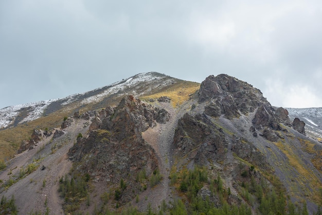 Colorful landscape with sunlit high green mountain with snow and sharp rocks under gray cloudy sky Dramatic view to large mountain with mossy top with snow under rainy clouds at changeable weather