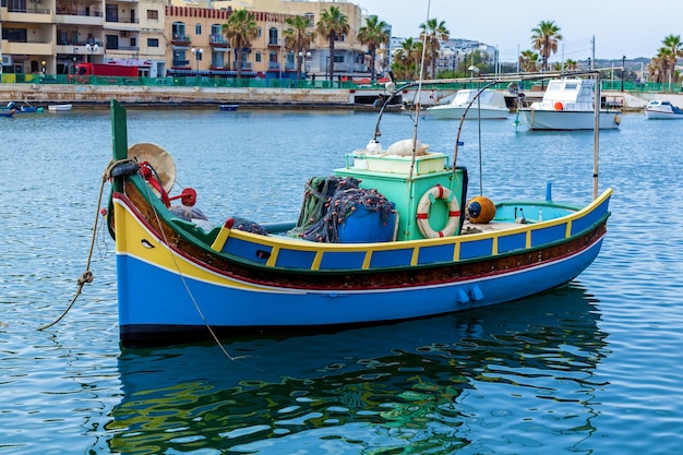 Красочная luzzu у берега рыбацкой деревни в солнечный день, Мальта. Рыболовная лодка в сине-желтых тонах находится в гавани.