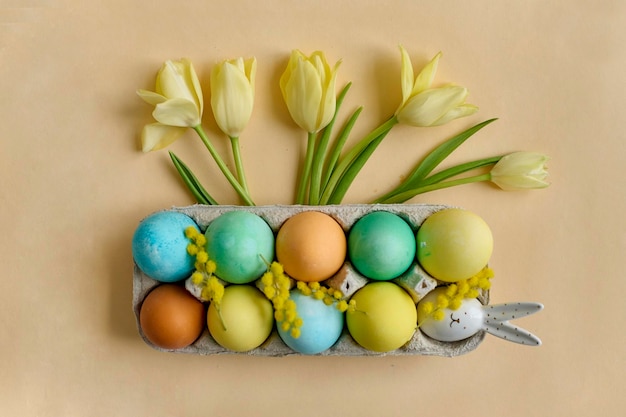 Colorful painted Easter eggs in a paper egg container with tulips on a beige background Top view