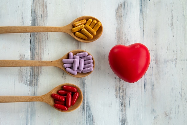 Colorful pills are in the spoon near rubber heart on white wooden table