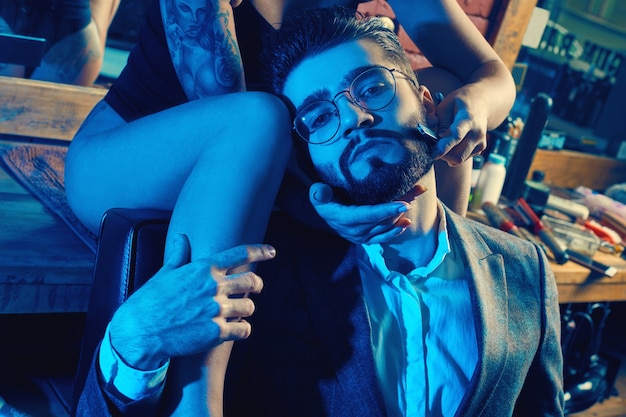 colorful portrait of beautiful couple: brutal man in elegant suit and sexy girl with a tattoo wearing lingerie in barbershop