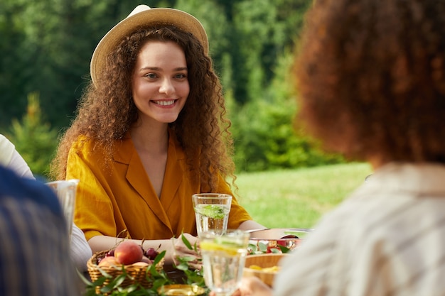 Colorful Portrait of smiling young woman enjoying dinner with friends outdoors while sitting at table during Summer party