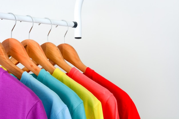 Magliette variopinte dell'arcobaleno che appendono sulla gruccia per vestiti di legno sopra fondo bianco