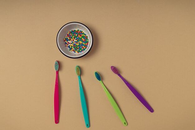 Colorful round ball sprinkles in a white small bowl on yellow paper background and four different colour vibrant toothbrushes. Creative dental care and healthy sugar consumption concept.
