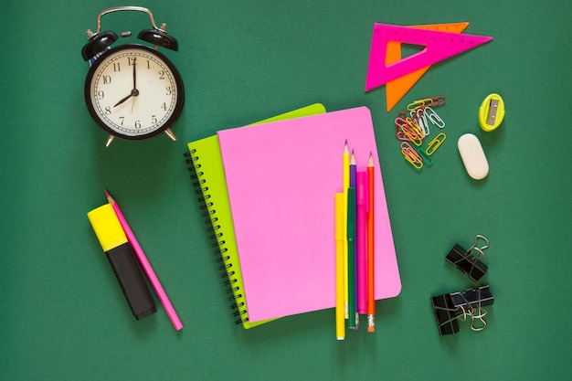 Photo colorful school supplies, pink book, and alarm clock on green.