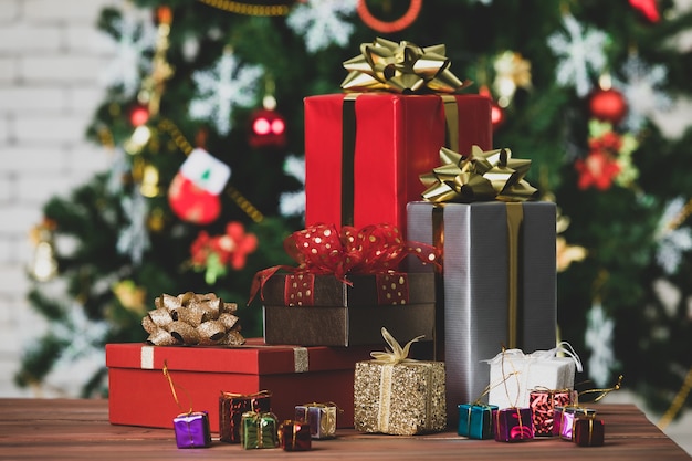 Colorful small and big present gift boxes with shiny ribbon bow\
tie placed on corner of wooden table in front fully decorative\
beautiful christmas eve pine tree and brick wall in blurred\
background.