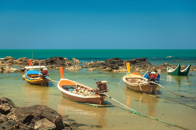 Colorful wooden Thai style fishing boats stand among the rocks against the backdrop of the sea and blue sky.