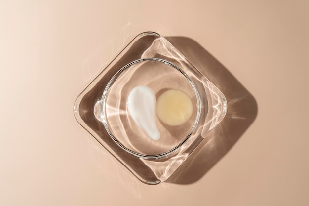 A colorful yellow drop of transparent face serum or essential oil and a smudge of a white face cream lying on a glass plate on a beige background