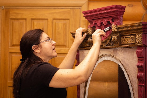 Coloring upper part of ornaments on shabby wooden cupboard in\
red color with big brush performed by engaged woman reuse of old\
workshop for old antique furniture restoration at home\
ecofriendly