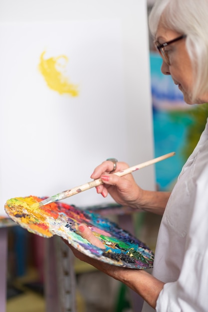 Colors and paint. Aged woman wearing white blouse holding color palette and painting with gouache