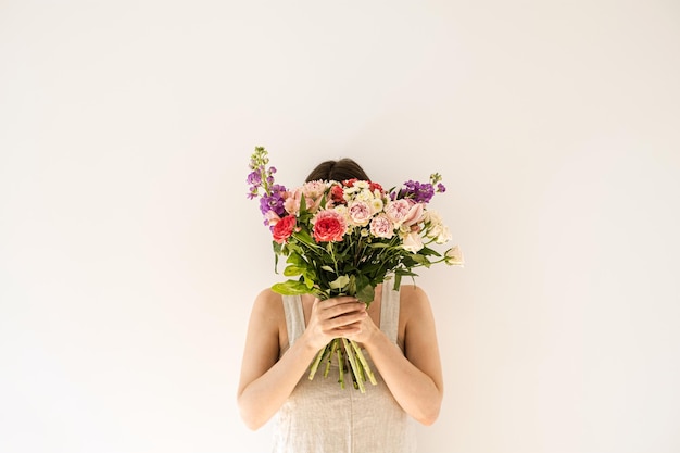 Colourful flowers bouquet in female hands on white background Young woman in neutral beige washed linen dress hide face Aesthetic beauty and fashion floral concept Face hidden by flower bouquet