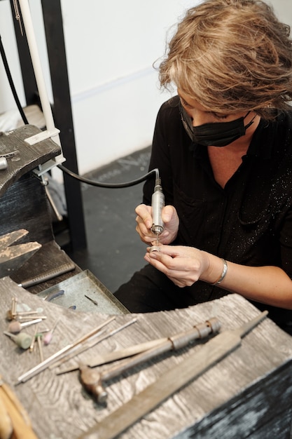 Comcentrated femele jeweler in mask sitting at work station and using abrasive nozzle while creating...