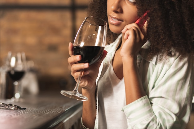 Venire. elegante giovane donna seduta al bancone del bar e bere vino mentre parla al telefono