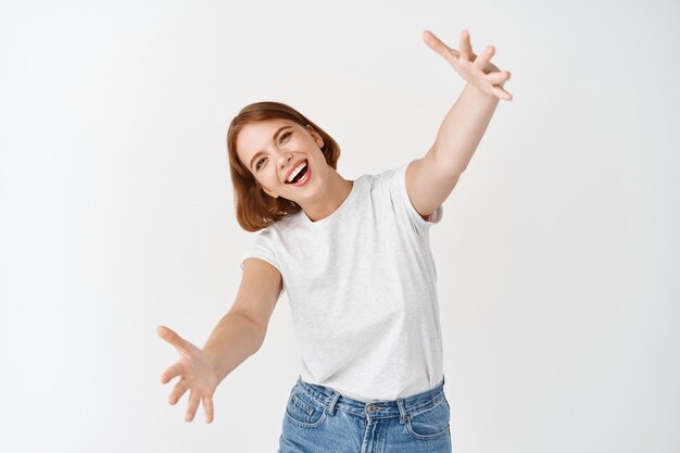 Come her for hug. Smiling happy woman stretch out hands for cuddle, welcome you, wants to embrace person, standing against white wall