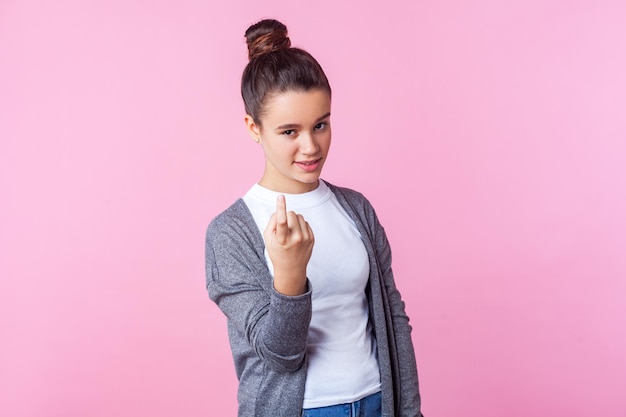 Photo come here portrait of beautiful brunette teenage girl with bun hairstyle in casual clothes looking playfully and beckoning to approach come closer hither gesture indoor studio shot pink background