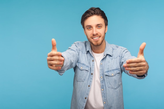 come into my arms. portrait of handsome friendly good-natured man in worker denim shirt raising hands to embrace, inviting for free hugs and smiling kind. studio shot isolated on blue background
